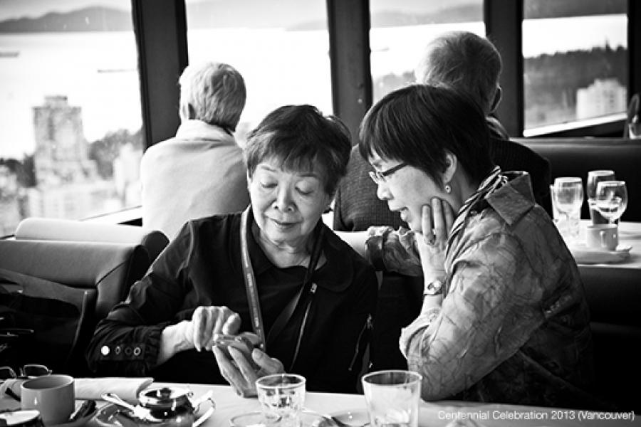 Two women sit at a dining table in a restaurant, engaged in conversation while examining a smartphone. Large windows behind them reveal a cityscape and body of water.