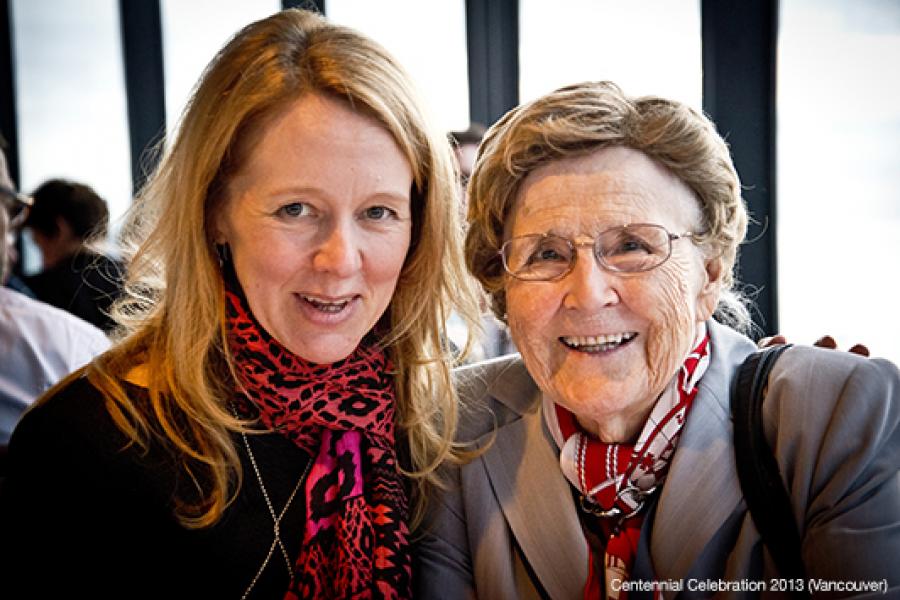 Two women smile warmly at the camera, one in a red and black scarf and the other in a light gray suit jacket, suggesting a close connection.