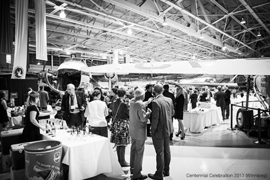 People gather and socialize at an event inside an aircraft hangar, surrounded by planes. Tables are set with drinks.People gather and socialize at an event inside an aircraft hangar, surrounded by planes. Tables are set with drinks.