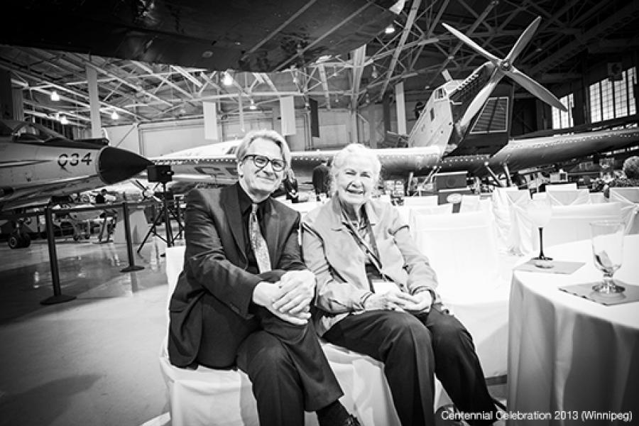People gather and socialize at an event inside an aircraft hangar, surrounded by planes. Tables are set with drinks.