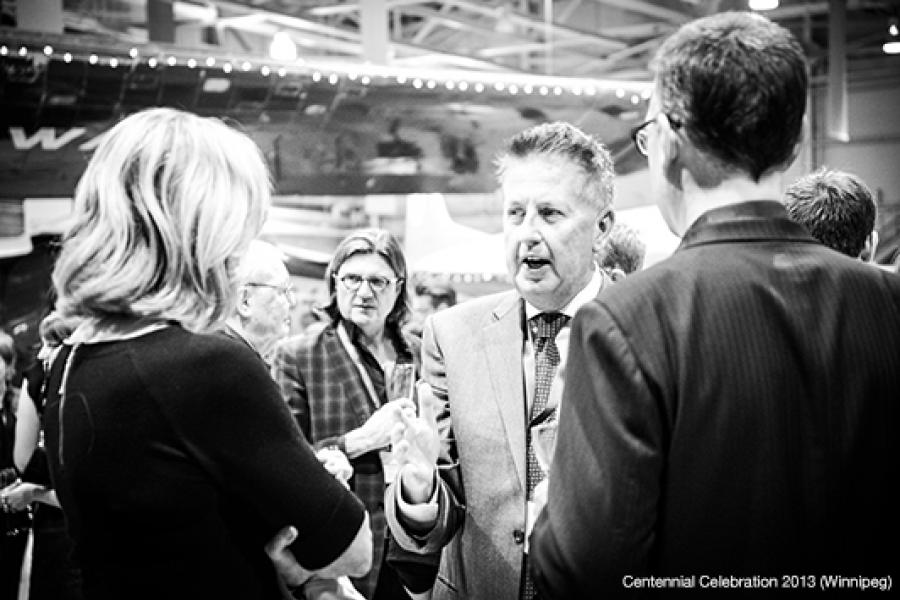 A group engages in conversation at an indoor event, with a man in a suit gesturing as he speaks, while others contribute to the lively atmosphere.