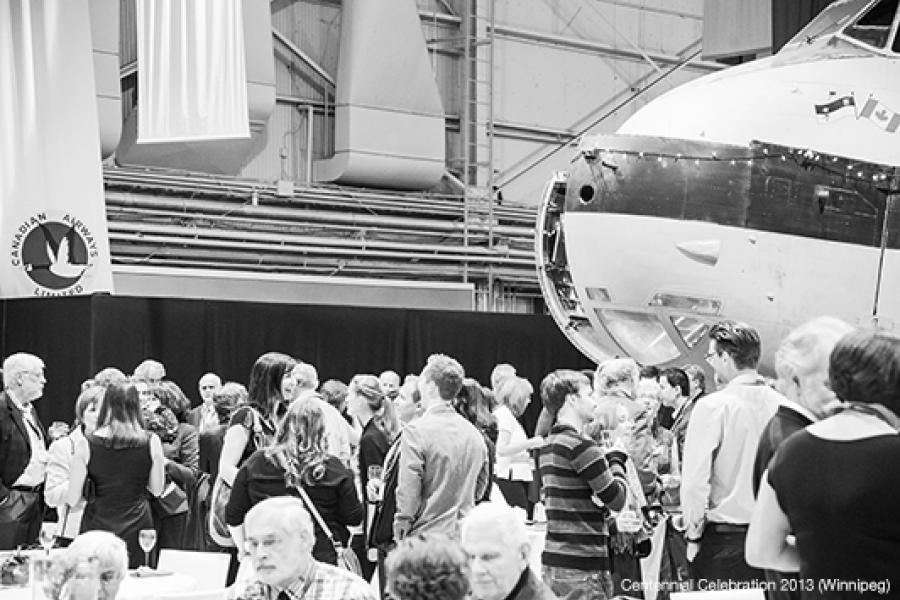 People gather and socialize at an event inside an aircraft hangar, surrounded by planes. 