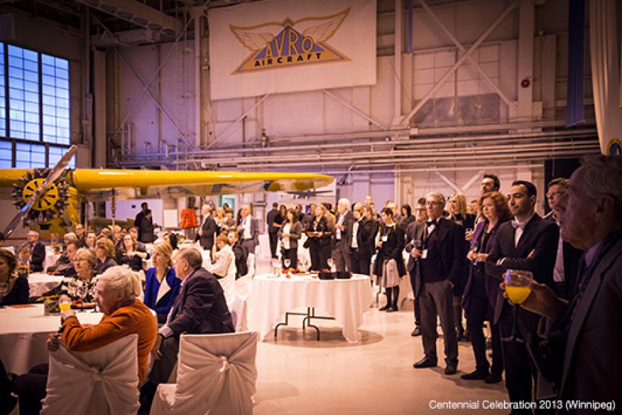  A large gathering in an aircraft hangar features people mingling around tables with white covers, under warm lighting and high ceilings.