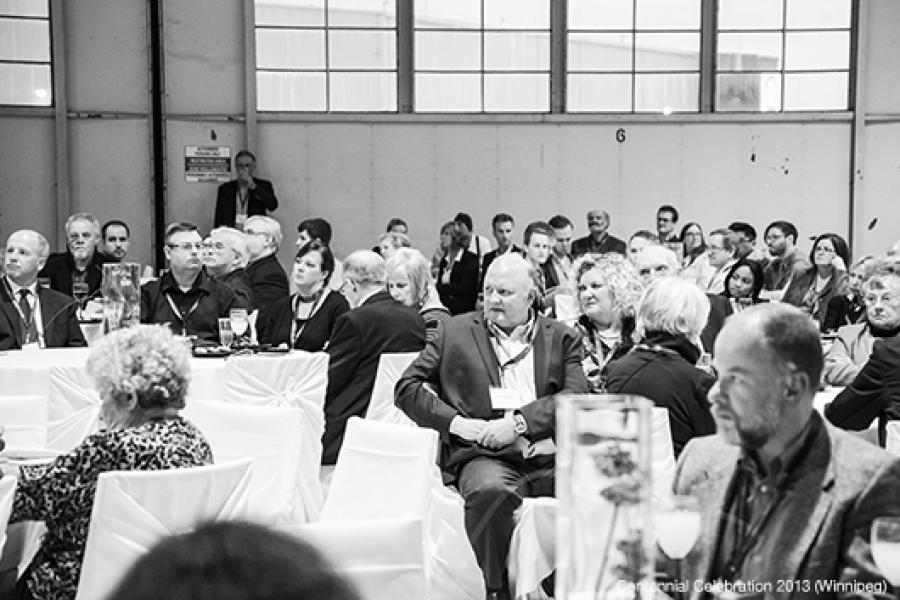 People gather and socialize at an event. Tables are set with drinks.