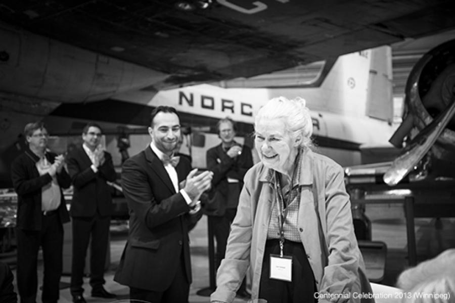 A celebratory event in front of a large aircraft features an elderly woman smiling warmly, with a joyful man clapping beside her, while others in formal attire also clap and smile, creating a cheerful atmosphere.