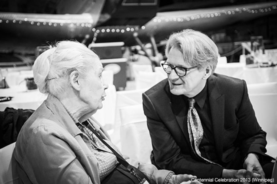 Two individuals engage in conversation at an aviation-themed indoor event, seated at a table in front of an aircraft. An elderly woman in a light jacket chats animatedly with a man in a dark suit and glasses.