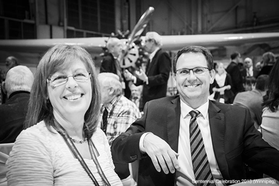 At a lively formal gathering, a man and woman smile at the camera while seated in front of a vintage airplane, with guests mingling in the blurred background.