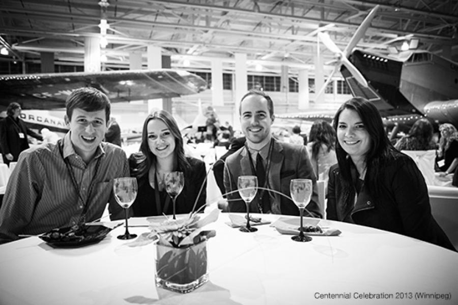 Inside an aircraft hangar, four smiling individuals in semi-formal attire sit around a circular table set with wine glasses and a small centerpiece, while large aircraft are visible in the background.