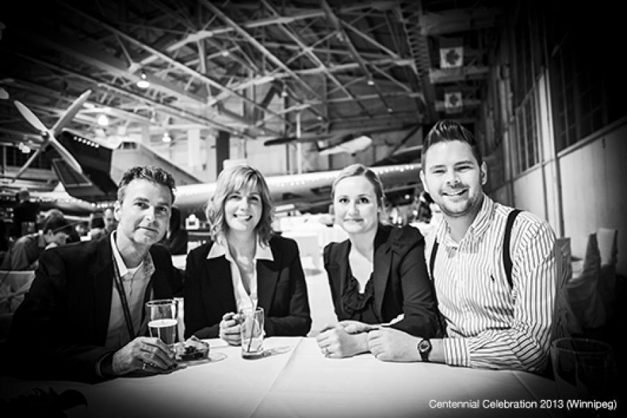 In a well-lit hangar, four people in formal attire smile at the camera while seated at a table with drinks, with a partially visible aircraft in the background, creating a celebratory aviation atmosphere.