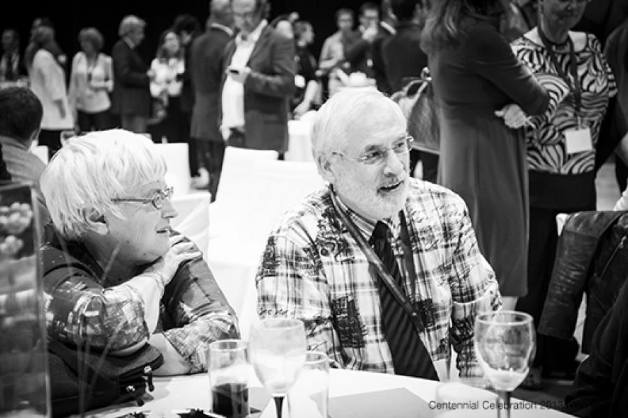 At a social gathering, two older adults—an engaged woman with short white hair and a man in a plaid shirt—sit at a table covered with a white tablecloth, surrounded by glassware. In the bustling background, other guests in professional attire are conversing.