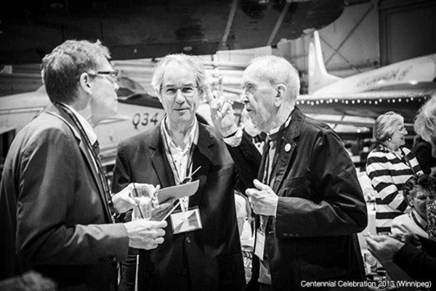 At a social gathering in an aviation-themed venue, three men in jackets engage in conversation, with visible aircraft parts in the hangar-like background, suggesting a formal or professional event.