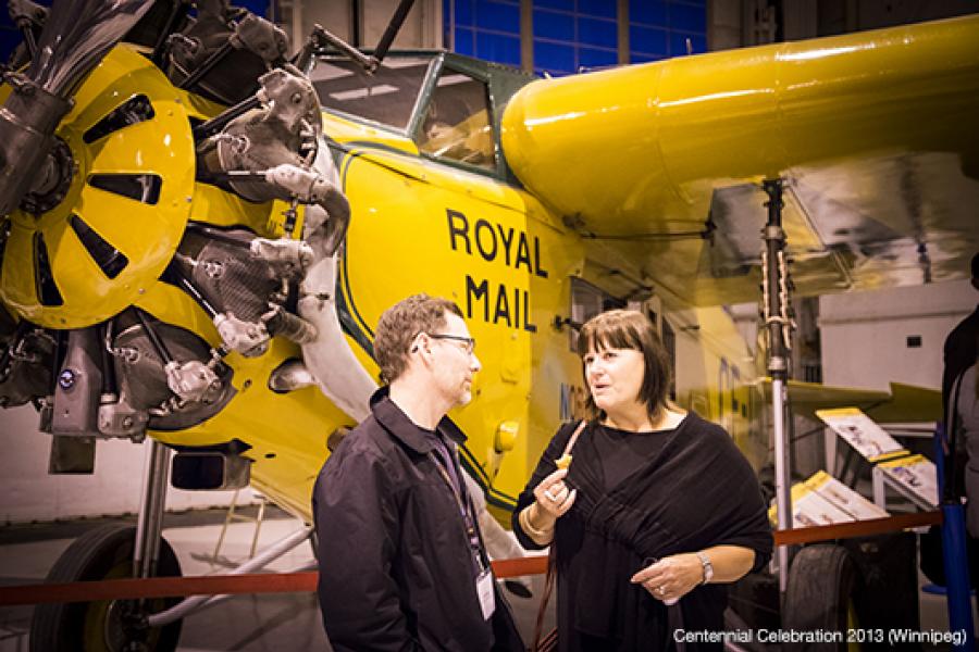 In an indoor exhibition featuring an old aircraft, a man and a woman engage in conversation in front of the radial engine. The warm lighting enhances the metallic finish of the aircraft, while information plaques and a red rope cordon are visible nearby.