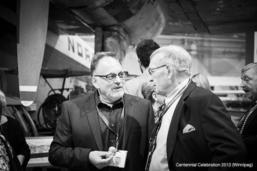 Two men in sharp suits engage in a lively conversation at an indoor event in an aircraft museum, surrounded by enthusiastic attendees.