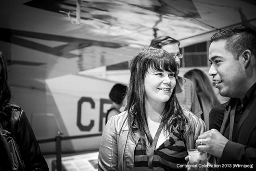 A woman and man, both smiling, engage in conversation amidst a bustling aviation-related event with soft lighting.