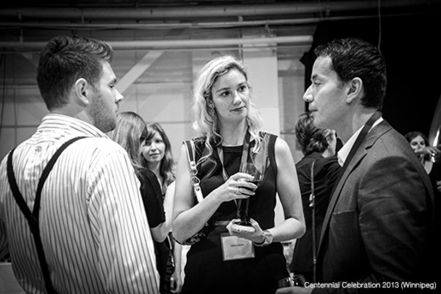 At an indoor event, three individuals converse, with others casually interacting in the background and structural elements above.
