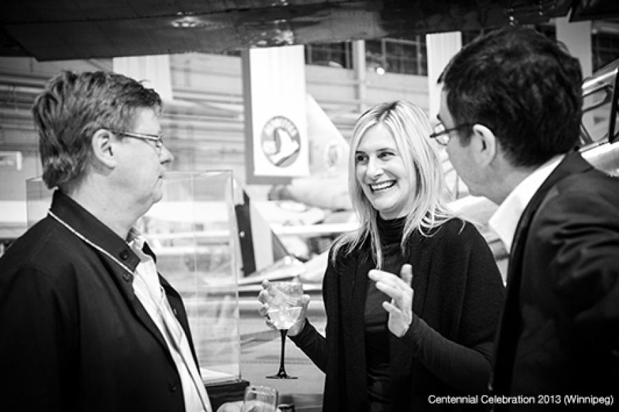 Three people engage in conversation at an indoor event, with aircraft exhibits and banners blurred in the background.