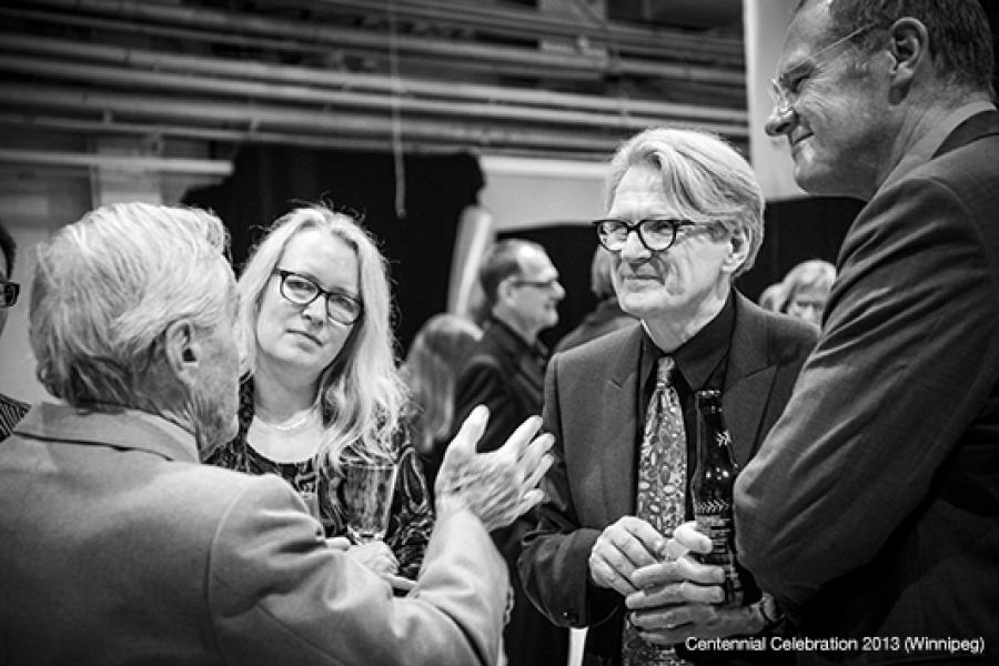 Four people engage in conversation at an event, with one gesturing, another listening seriously, and two in suits smiling.