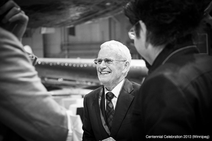 An elderly man in a suit smiles broadly while conversing with two suited individuals at an indoor celebration.