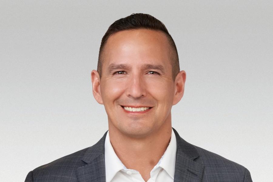 Smiling person in gray checkered suit jacket and white shirt against a gray background.