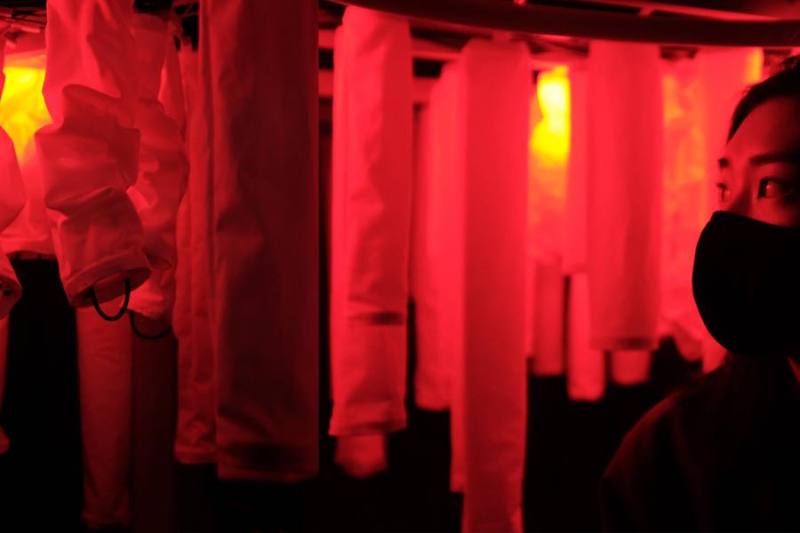 A person with a face mask stands in a room with red lighting and hanging white fabrics.