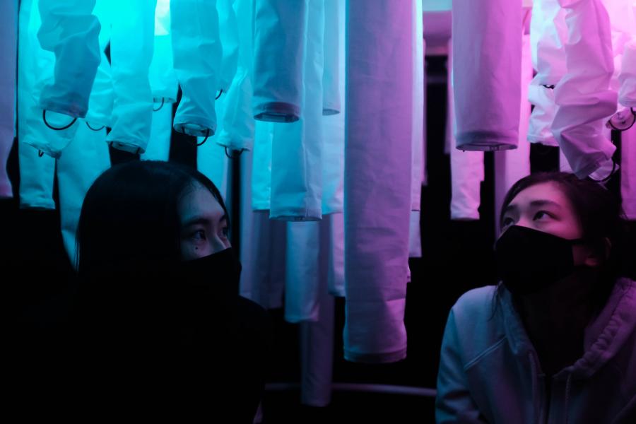 Two individuals wearing masks look up at illuminated hanging fabrics in shades of blue and purple.