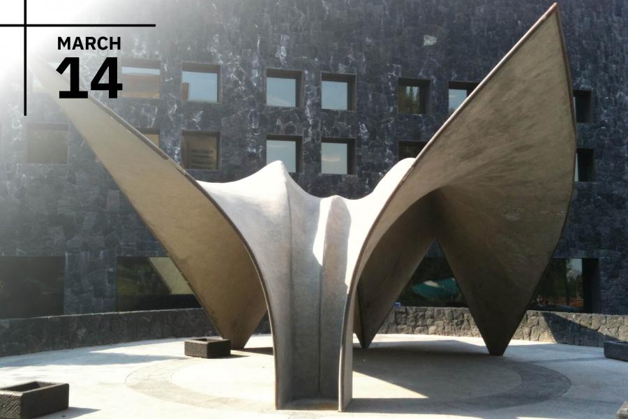 Abstract concrete sculpture with wing-like forms in front of a dark stone building.