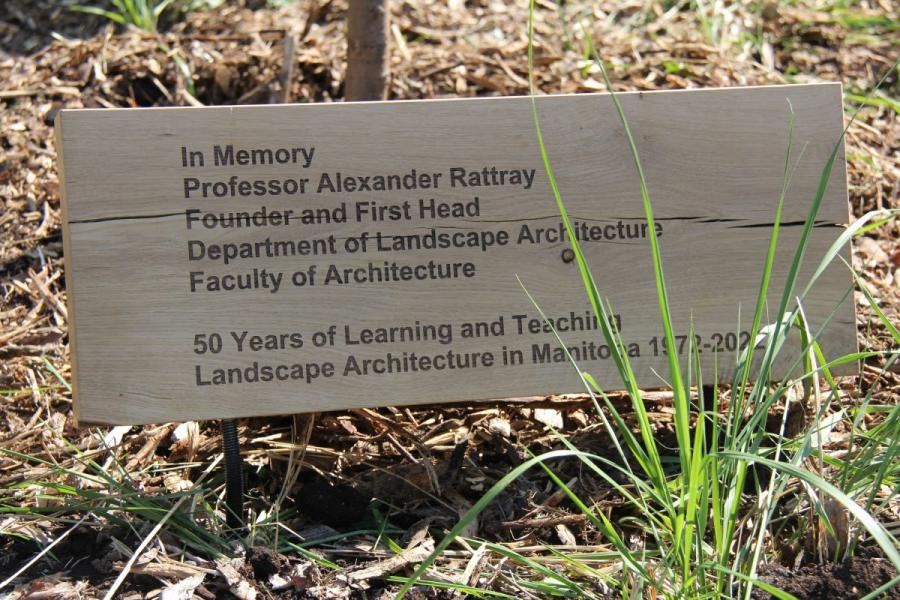 A wooden memorial plaque in a garden setting honors Professor Alexander Rattray, founder of Landscape Architecture at Manitoba, celebrating 50 years.