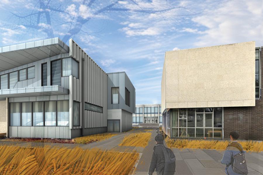 Modern architecture featuring a gray, industrial building with large windows and sharp angles. Two people walk along a path with dry grass under a blue sky.