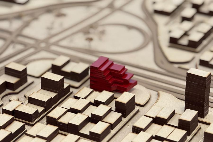 Architectural model featuring rectangular beige and brown buildings with a unique, stepped red structure. Background shows roads, suggesting urban planning.