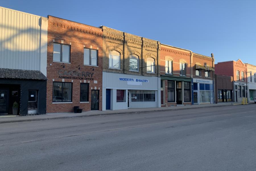 Street view of a row of historic brick buildings under a clear blue sky. Signs include "Carberry Administration Office" and "Modern Bakery." Quiet, small-town ambiance.