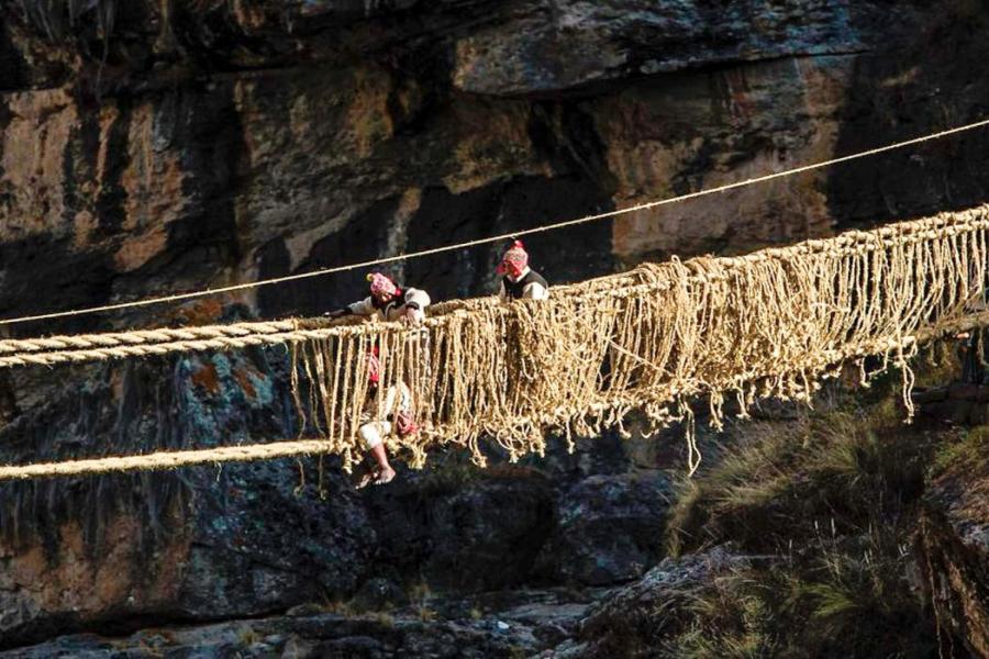A group of people rebuilds a traditional rope bridge over a rocky gorge. The scene conveys teamwork and cultural heritage against a rugged backdrop.