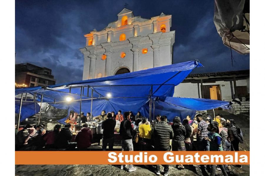 Nighttime scene of people gathering under blue tarps in front of a lit white church. The sky is dark and the mood is lively and communal.