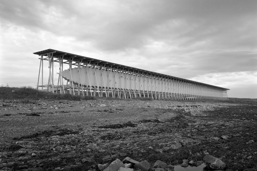 Black-and-white photo of a long, wooden fish drying rack on a rocky coastline under a cloudy sky, conveying a stark, industrial feel.