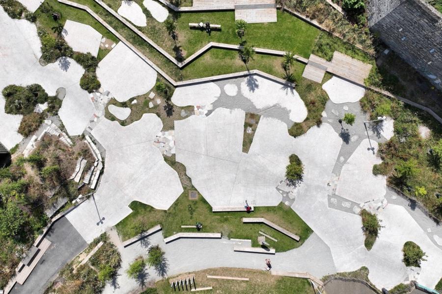 Aerial view of a park with geometric paths, grass, and scattered trees. Concrete walkways create a modern, abstract design. People appear small, adding scale.