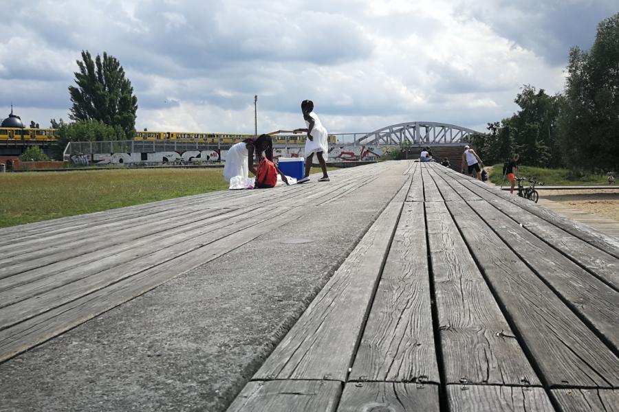 Wide-angle view of a boardwalk with people in casual attire sitting and walking. A yellow train and a bridge are visible under a cloudy sky. Calm atmosphere.