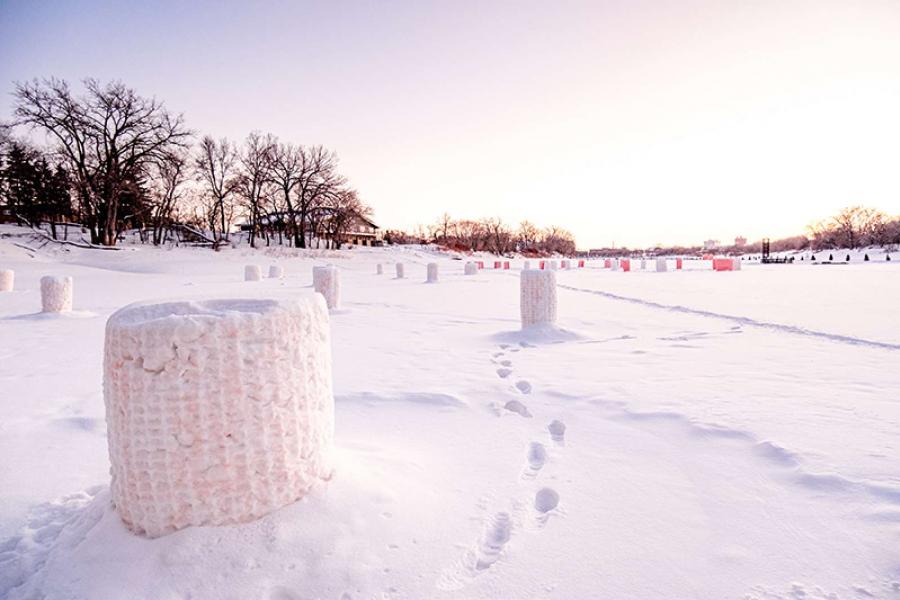 Snow-covered park at sunset with rows of cylindrical ice sculptures, footprints leading away, and bare trees in the distance under a pinkish sky.