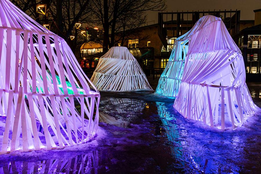 Glowing, sculptural structures made of white ribbons illuminate with purple and blue lights in a snowy outdoor setting, evoking a mystical, serene atmosphere.