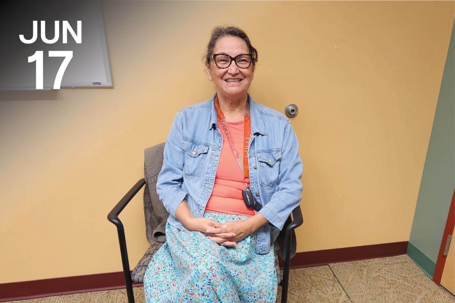 Grandmothers sitting on an office chair smiles infront of the camera as she wears a bright blue skirt and denim button up top.