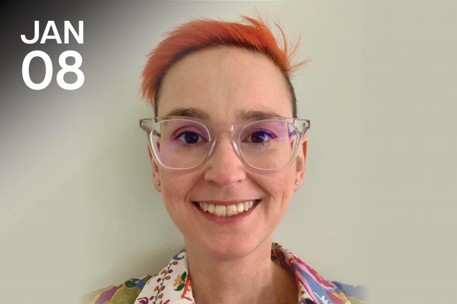A person with short red hair and clear glasses smiles warmly. They wear a colorful shirt with a floral pattern. "JAN 08" is displayed in the upper left corner.