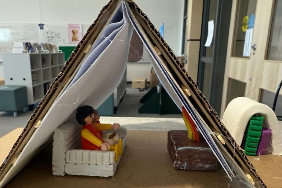 A cardboard and paper pyramid houses a toy figure sitting in a white armchair. A small campfire is nearby. The scene is set in a spacious room.