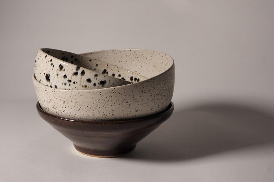Three stacked ceramic bowls in earthy tones; top bowl is speckled white with black spots, middle is speckled grey, bottom is dark brown. Soft, minimalist lighting.