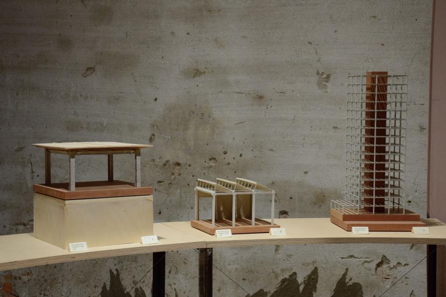 Three architectural models on display: a pagoda, a modern structure, and a high-rise building. They rest on a curved wooden table against a textured gray wall.