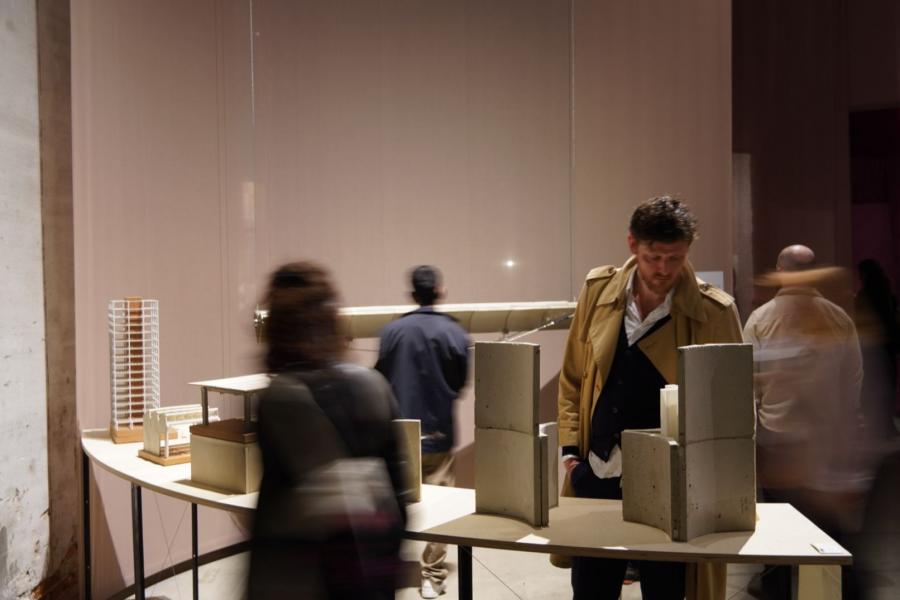 A man in a tan coat observes architectural models on a table in a dimly lit exhibit. Blurred figures suggest movement and add a dynamic, contemplative mood.