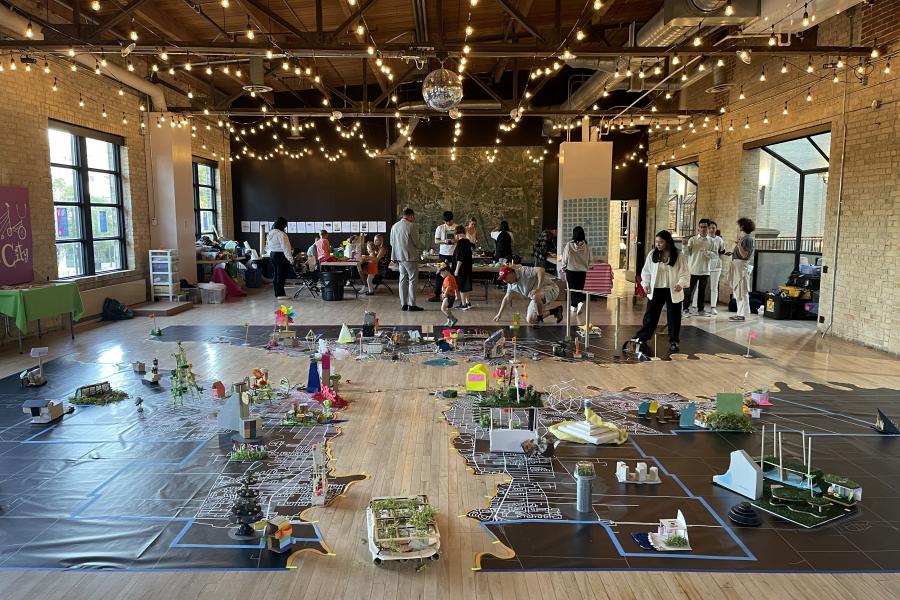 A spacious room with high ceilings and string lights showcasing a large floor map with colorful models. People gather around, engaging in a creative, collaborative atmosphere.