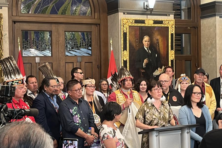 A diverse group of people, some in traditional attire, stand at a podium in a formal setting with flags and a large historical portrait behind them. 