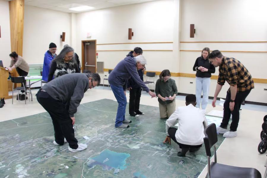 A group of nine people gathers around a large map placed on the floor of a brightly lit room, actively discussing and pointing, suggesting collaboration.