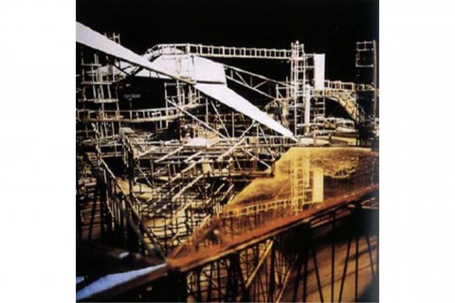 Nighttime industrial construction site with dense steel scaffolding, platforms, beams, and a large diagonal conveyor structure; rugged, dramatic atmosphere.