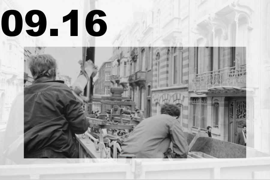 Black-and-white street scene: two men ride in an open truck past ornate historic buildings, evoking a busy, gritty urban atmosphere.