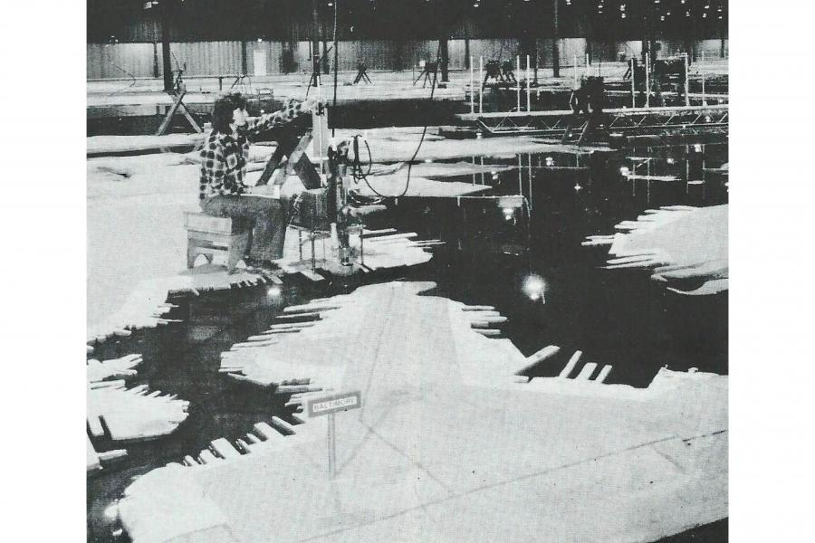 Black-and-white outdoor scene with people gathered beside a dark, reflective pond, surrounded by snow and jagged shapes resembling piano keys.