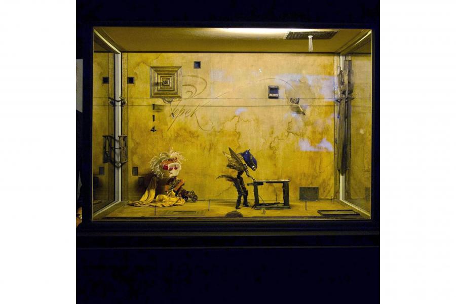 A yellowed, graffiti-marked room in a glass display case, featuring a seated puppet and a dark birdlike figure beside a small table.